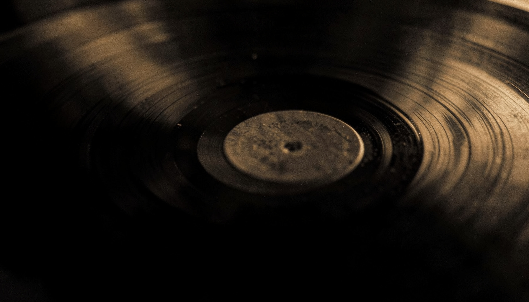 A close-up duotone photograph of a vinyl record groove catching light, rendered in deep charcoal black and warm amber tones.