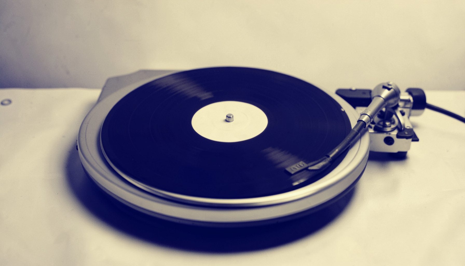 A warm duotone photograph of a vinyl record on a turntable platter, rendered in deep indigo and cream tones, with soft ambient light