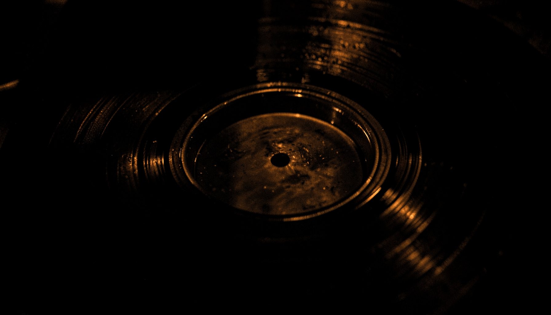A dark, moody close-up of a vinyl record groove catching low amber light, deep blacks and warm copper highlights.